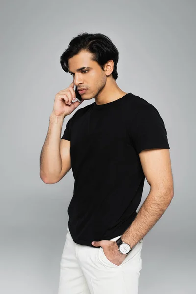 Молодий чоловік розмовляє на мобільному телефоні і позує рукою в кишені ізольовано на сірому — стокове фото