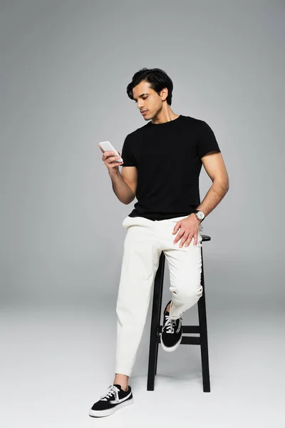 Full length of young man using cellphone while leaning on chair on grey — Stock Photo