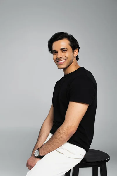 Cheerful young man smiling and sitting on chair isolated on grey — Stock Photo