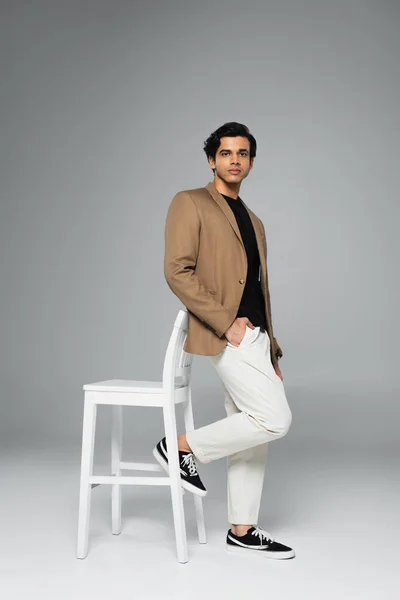 Full length of young man in blazer posing with hand in pocket near white chair on grey — Stock Photo