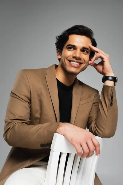 Happy young man in blazer sitting on white chair isolated on grey — Stock Photo