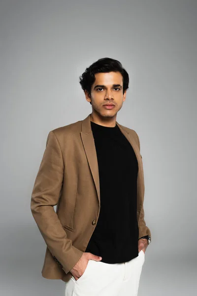 Young man in blazer posing with hands in pockets isolated on grey — Stock Photo