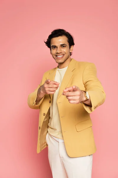 Joven feliz en chaqueta amarilla señalando con los dedos y mirando a la cámara en rosa - foto de stock