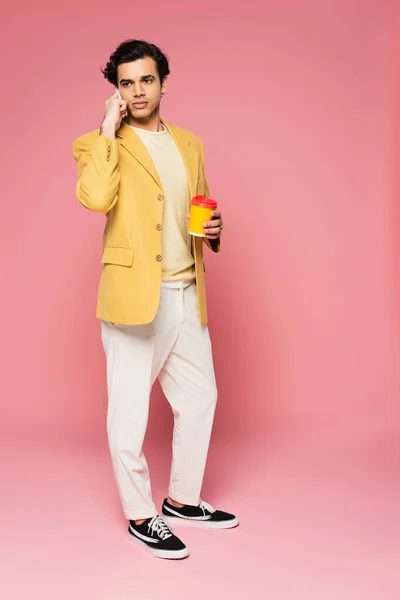 Longitud completa de hombre joven hablando en el teléfono celular y sosteniendo la taza de papel en rosa - foto de stock