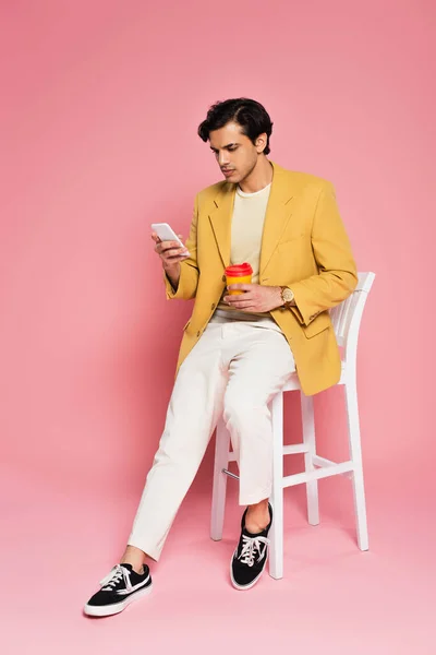 Pleine longueur de jeune homme utilisant un téléphone portable et tenant tasse en papier tout en étant assis sur une chaise blanche sur rose — Photo de stock