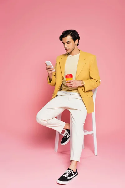 Full length of young man using smartphone and holding paper cup while sitting on white chair on pink — Stock Photo