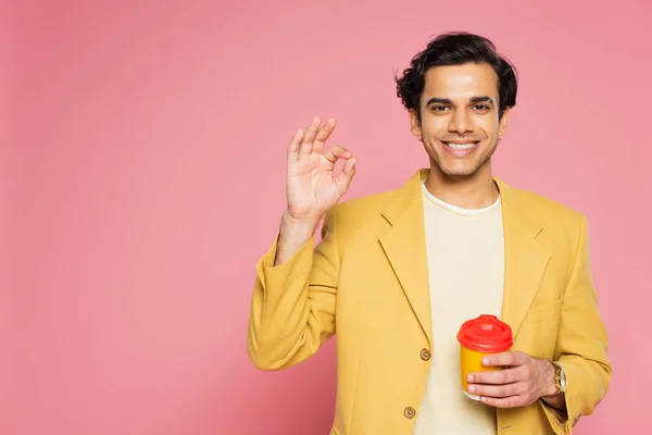 Щасливий молодий чоловік в жовтому блістері, показуючи знак ОК і тримаючи паперову чашку ізольовано на рожевому — стокове фото