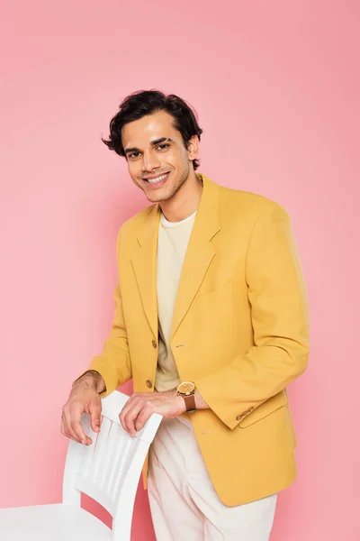 Jovem feliz em blazer amarelo encostado na cadeira branca isolado em rosa — Fotografia de Stock