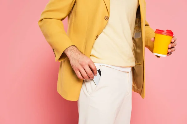 Cropped view of young man holding paper cup while putting credit card in pocket on pink — Stock Photo