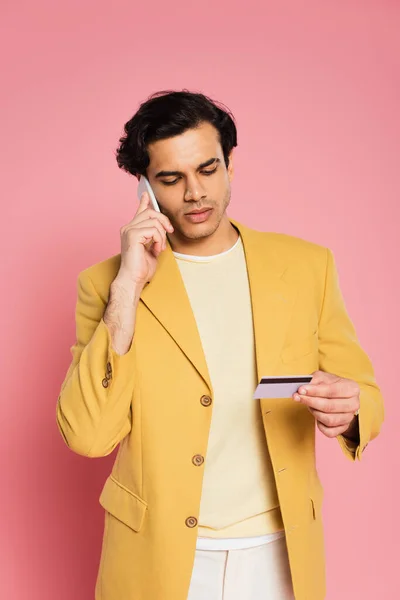 Hombre en chaqueta amarilla con tarjeta de crédito mientras habla en el teléfono inteligente aislado en rosa - foto de stock