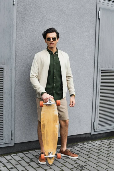 Piena lunghezza di uomo elegante in occhiali da sole tenendo longboard vicino edificio — Foto stock