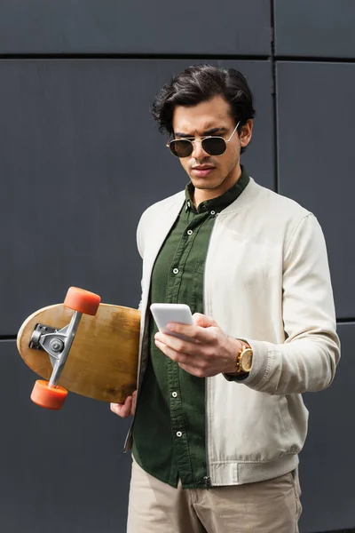 Élégant jeune homme en lunettes de soleil et blouson de bombardier tenant longboard tout en textant sur smartphone près du bâtiment — Photo de stock