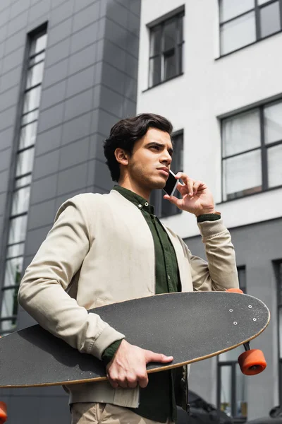 Stilvoller junger Mann in Bomberjacke hält Longboard in der Hand, während er in der Nähe des Gebäudes mit dem Smartphone spricht — Stockfoto