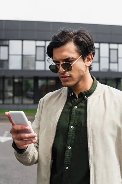 Jovem elegante em óculos de sol e jaqueta bomber mensagens de texto no smartphone perto do edifício — Fotografia de Stock