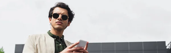 Joven con estilo en gafas de sol y chaqueta de bombardero celebración de teléfono inteligente en el exterior, bandera - foto de stock