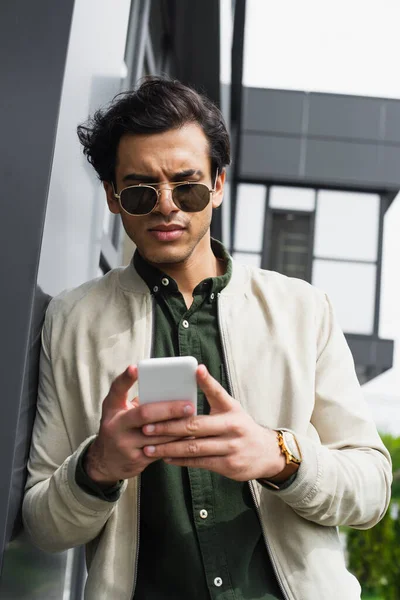 Jovem elegante em óculos de sol e casaco de bombista segurando telefone celular perto do edifício — Fotografia de Stock