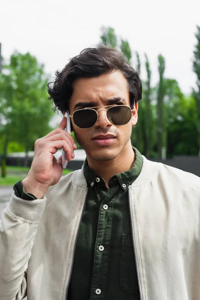 Élégant jeune homme en lunettes de soleil et blouson de bombardier parler sur téléphone mobile — Photo de stock