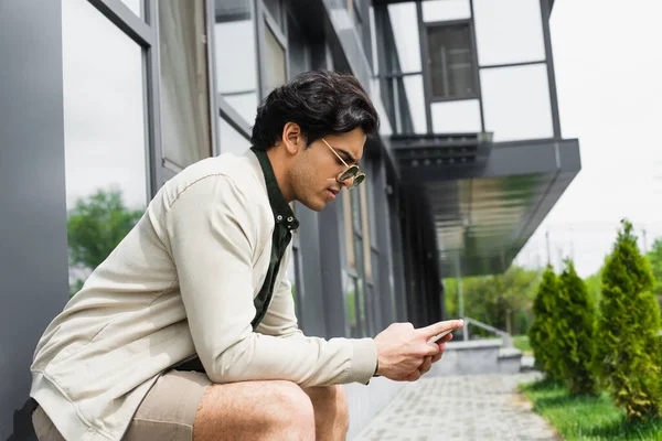 Vue latérale du jeune homme élégant dans les lunettes de soleil bavarder sur le téléphone mobile près du bâtiment — Photo de stock