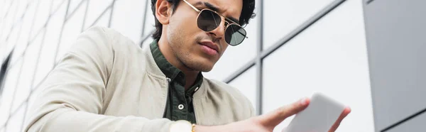 Стильний молодий чоловік в сонцезахисних окулярах спілкується на мобільному телефоні біля будівлі, банер — стокове фото