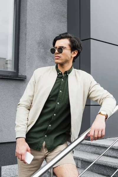 Joven con estilo en gafas de sol sosteniendo el teléfono móvil mientras posa cerca del edificio - foto de stock