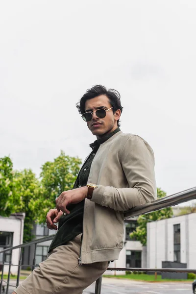 Young man in stylish sunglasses and bomber jacket posing outside — Stock Photo