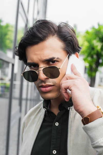 Joven con estilo en gafas de sol hablando en el teléfono móvil y mirando a la cámara - foto de stock