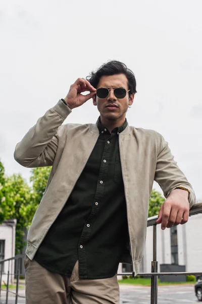 Young man in bomber jacket adjusting stylish sunglasses while posing outside — Stock Photo