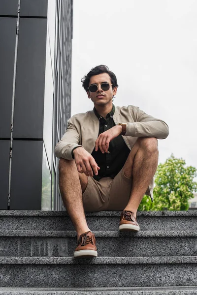 Full length of young man in stylish sunglasses and bomber jacket sitting on stairs near building — Stock Photo