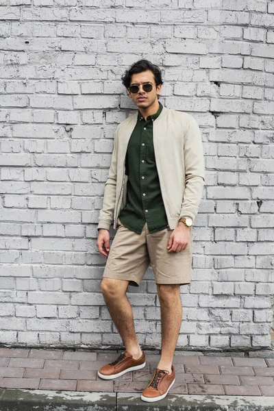 Pleine longueur de jeune homme dans des lunettes de soleil élégantes et une veste de bombardier posant près du mur de briques — Photo de stock