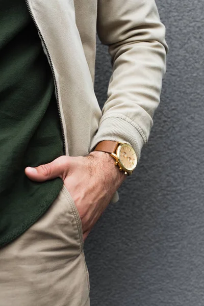 Vista recortada de hombre joven con elegante reloj de pulsera posando con la mano en el bolsillo cerca de la pared gris - foto de stock