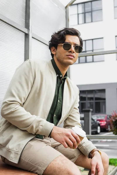 Young man in stylish sunglasses and bomber jacket holding coffee to go outside — Stock Photo