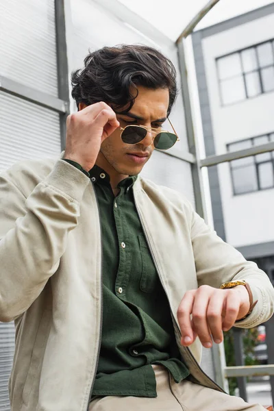 Junger Mann passt stylische Sonnenbrille an, während er auf Armbanduhr schaut — Stockfoto