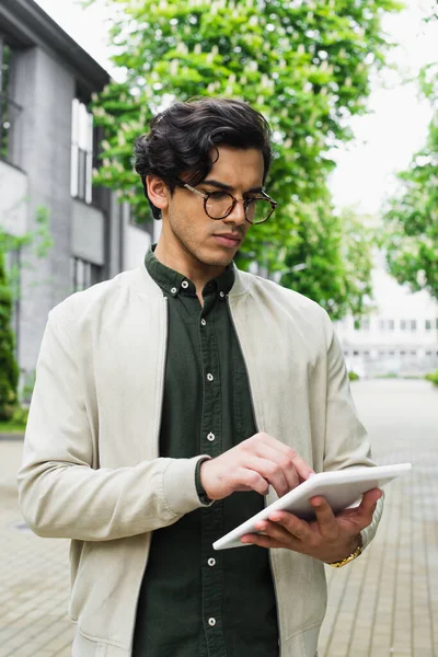 Young man in eyeglasses and bomber jacket using digital tablet outside — Stock Photo