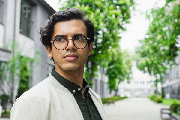 Young man in eyeglasses and bomber jacket looking away outside — Stock Photo