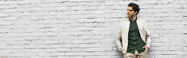 Молодой человек в куртке бомбардировщика, стоящий с руками в карманах возле кирпичной стены, баннер — стоковое фото