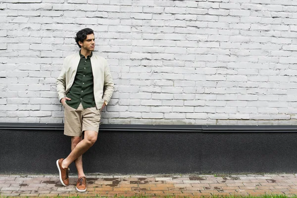 Full length of young man in bomber jacket standing with hands in pockets near brick wall — Stock Photo