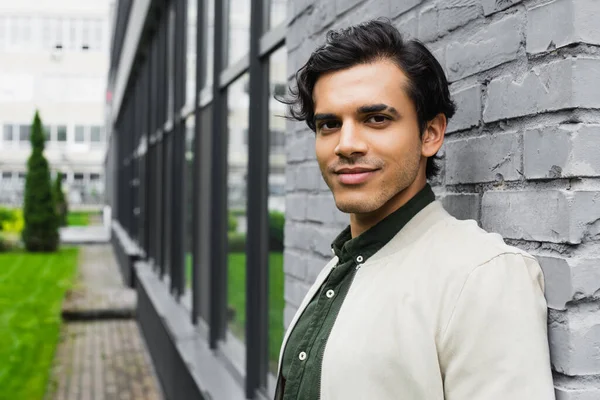 Cheerful young man in bomber jacket smiling near brick wall — Stock Photo