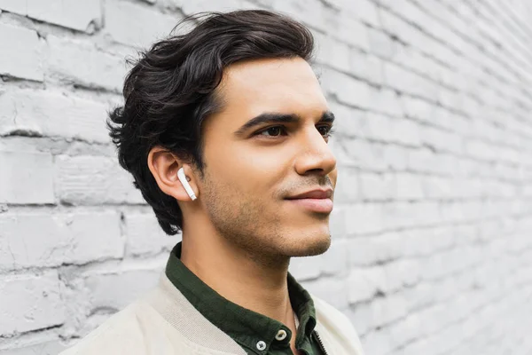 Cheerful young man in wireless earphones smiling while listening music near brick wall — Stock Photo