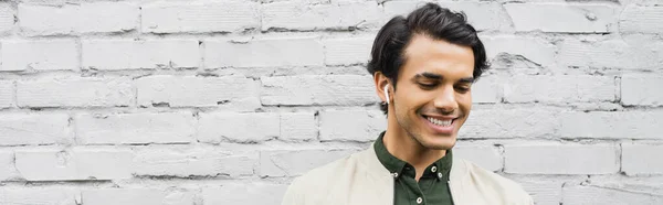 Fröhlicher junger Mann mit drahtlosen Kopfhörern, der lächelt, während er Musik in der Nähe von Ziegelmauer hört, Banner — Stockfoto