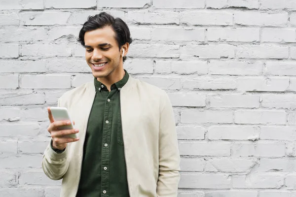 Cheerful young man in wireless earphones smiling while using smartphone near brick wall — Stock Photo