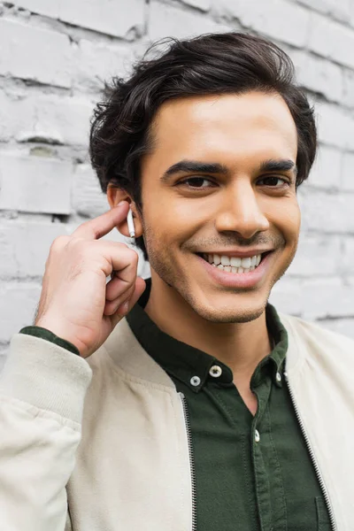 Cheerful young man adjusting wireless earphones and smiling near brick wall — Stock Photo