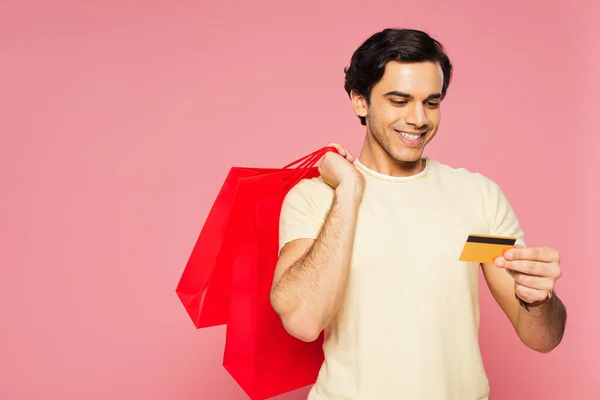 Jovem feliz segurando sacos de compras vermelhos enquanto olha para o cartão de crédito isolado em rosa — Fotografia de Stock