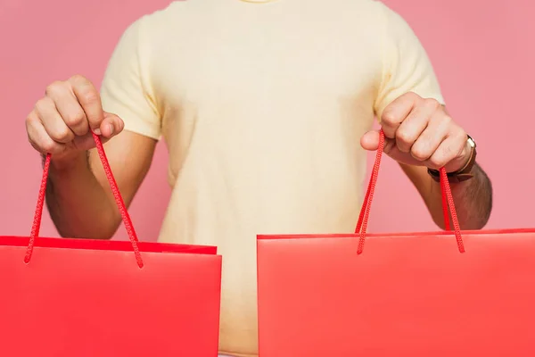 Vista cortada de jovem segurando sacos de compras vermelhos isolados em rosa — Fotografia de Stock