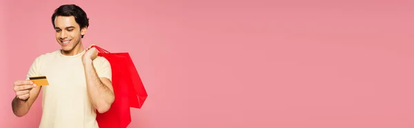 Jovem feliz segurando sacos de compras vermelhos enquanto olha para o cartão de crédito isolado em rosa, banner — Fotografia de Stock