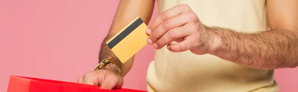 Vista recortada del joven que pone la tarjeta de crédito en el bolso rojo de la compra aislado en rosa, bandera - foto de stock