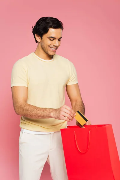 Allegro giovane che mette la carta di credito nella shopping bag rossa isolata su rosa — Foto stock