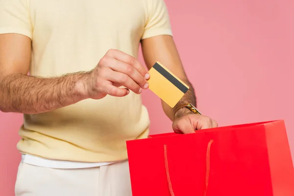 Vista recortada de un joven que pone la tarjeta de crédito en una bolsa roja aislada en rosa - foto de stock