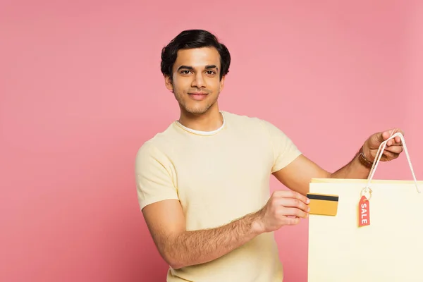Fröhlicher junger Mann mit Kreditkarte und Einkaufstasche mit Verkaufsanhänger isoliert auf rosa — Stockfoto