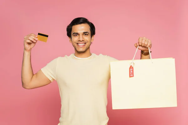 Smiling young man holding credit card and shopping bag with sale tag isolated on pink — Stock Photo