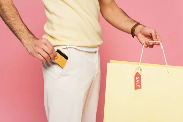 Обрізаний вид молодого чоловіка, що кладе кредитну картку в кишеню і тримає сумку з торговою маркою ізольовано на рожевому — стокове фото